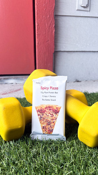 Spicy Pizza flavor protein bar propped up next to yellow dumbbells on green gym turf grass.