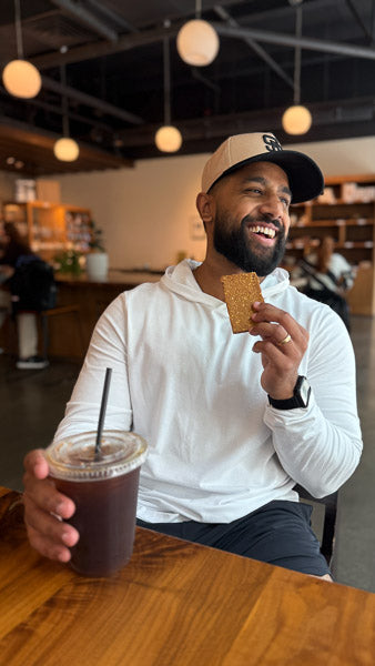 Man at a coffee shop table holding an iced coffee and enjoying a protein bar.