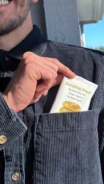 Hand placing an Everything Bagel flavor protein bar into a shirt pocket for a convenient portable snack.