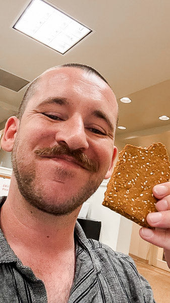 Man with a mustache smiling while holding an unwrapped, savory protein bar that highlights sesame seeds.