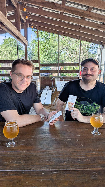 Two men sitting at a outdoor patio table pairing a savory protein bar with glasses of beer.