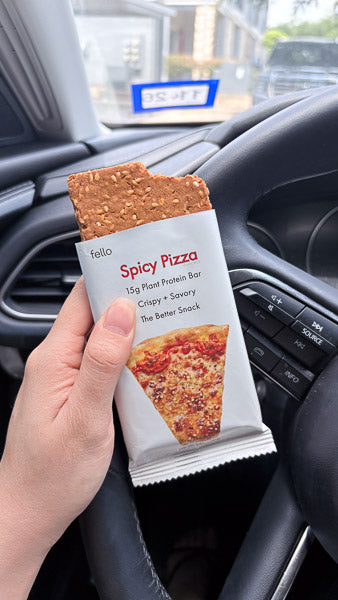 Hand holding a Spicy Pizza flavor protein bar over a steering wheel for a road trip car snack.