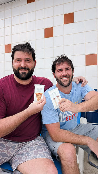 Two friends laughing and holding up high-protein snack bars outside.