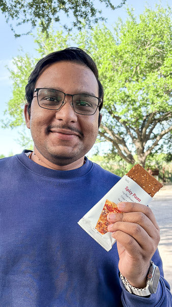 Man with glasses outdoors holding a packaged Spicy Pizza flavor savory protein bar.