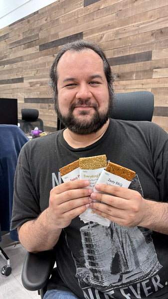 Man in an office chair holding a variety pack sample of three savory plant-based protein bars.
