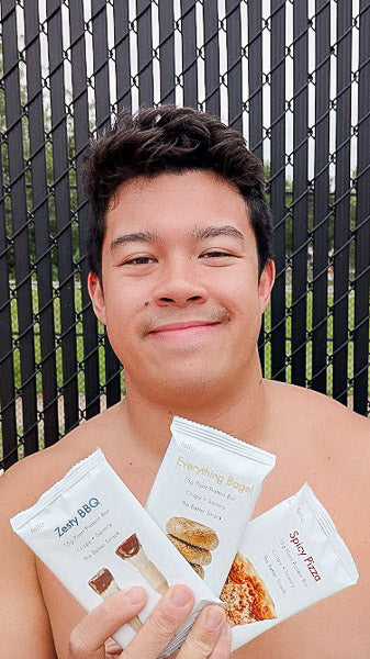 Smiling man outdoors holding a collection of savory protein bars including Everything Bagel and Zesty BBQ.