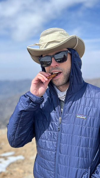 Man wearing a sun hat and jacket eating an energy protein bar while hiking a mountain trail.