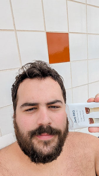 Bearded man holding up a Zesty BBQ flavor protein bar against a white and brown tiled wall backdrop.