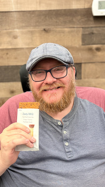 Smiling man wearing a flat cap and glasses holding up a nutritious savory protein bar.
