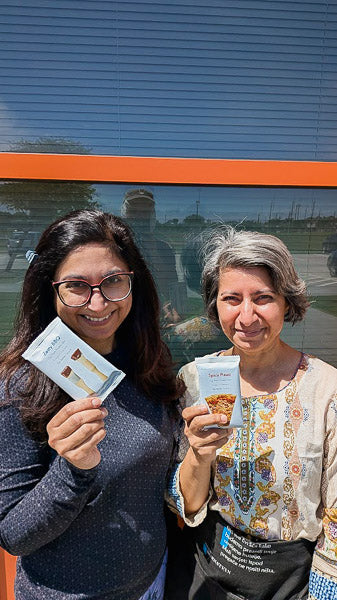 Two smiling women standing outside holding packaged savory plant-based protein bars.