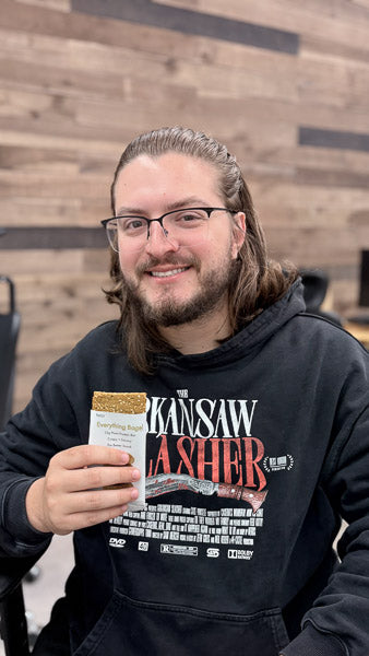 Man in a black graphic hoodie holding a protein bar in front of a wooden wall backdrop.