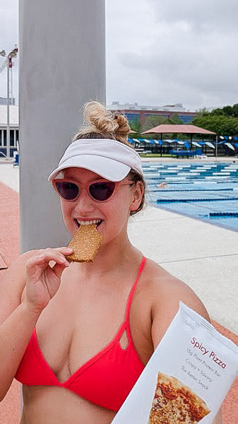 Woman in a red swimsuit by a swimming pool eating a Spicy Pizza flavor protein bar.