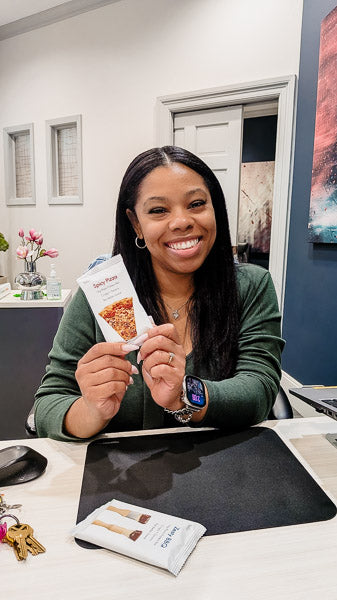 Smiling woman holding a protein bar at her office desk for a midday workday snack.