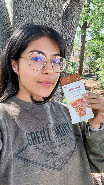 Woman outdoors wearing glasses and holding up a Spicy Pizza flavor protein bar.