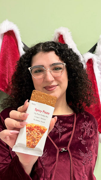 Woman with glasses holding a Spicy Pizza flavor protein bar with festive holiday decorations in the background.
