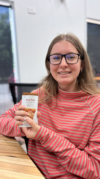 Woman smiling at a kitchen table holding a nutritious protein bar.